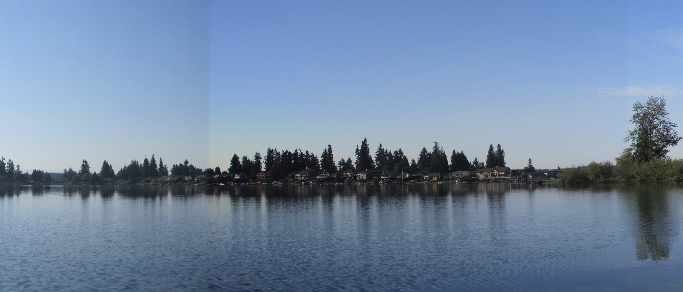 Wide view across the still water of Lake Tapps toward a tree-lined shore dotted with homes among the firs, their reflections mirrored in the calm surface under a clear daytime sky